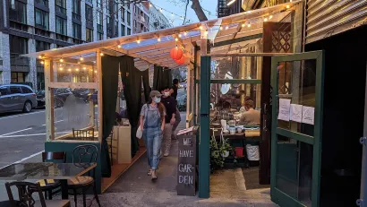 Person wearing mask walking through temporary outdoor dining setup lined with bistro lights at dusk in New York City.
